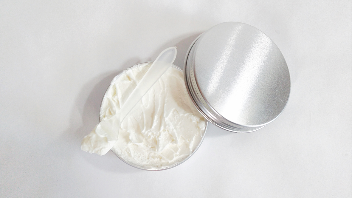 Hydratačné telové maslo v kovovej nádobke s priehľadnou plastovou lyžičkou naberajúcou hmotu.